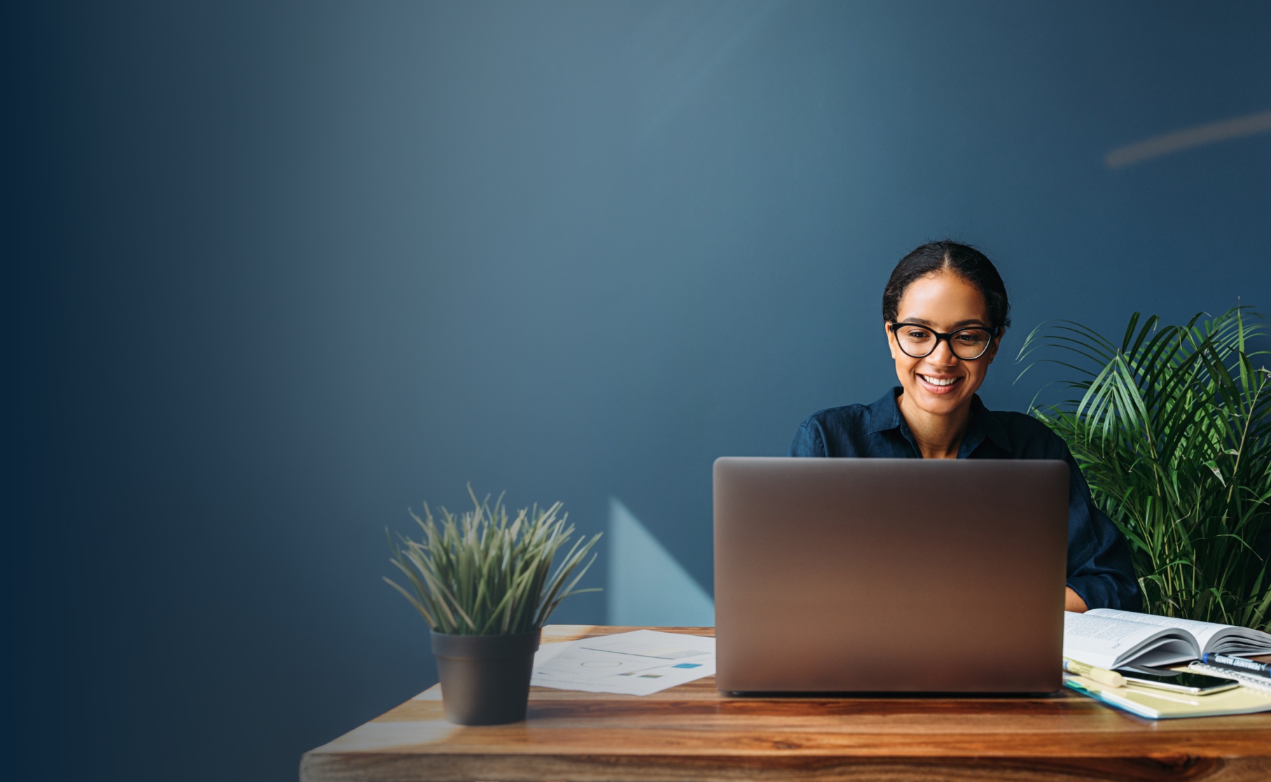 A woman seated at a desk, focused on her laptop, with a notepad and pen beside her